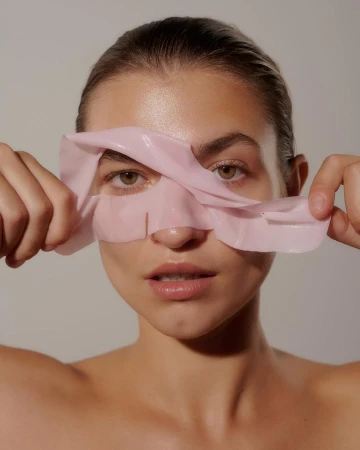Close-up of a woman removing a pink sheet mask from her face, looking directly at the camera against a neutral background.