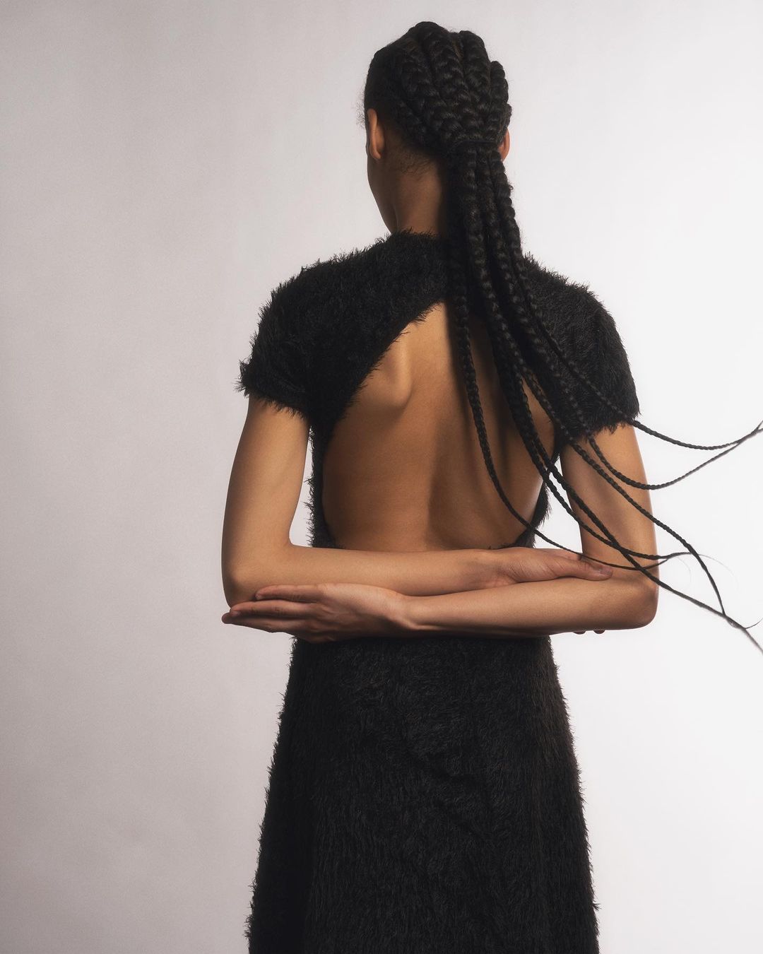 Back view of a woman with cornrow braids wearing a black, open-back dress in a studio setting, showcasing fashion and style.