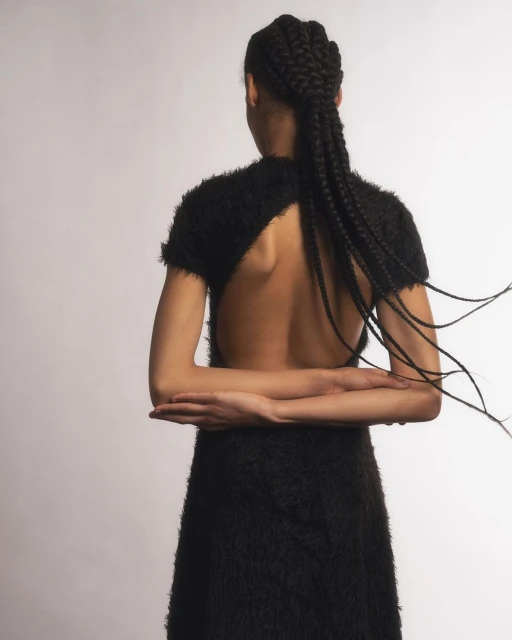 Back view of a woman with cornrow braids wearing a black, open-back dress in a studio setting, showcasing fashion and style.