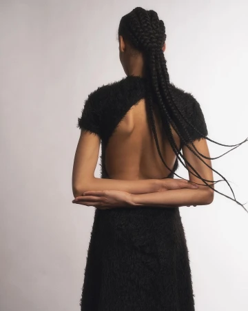 Model seen from behind wearing a textured black dress with long braided hair against a plain white background