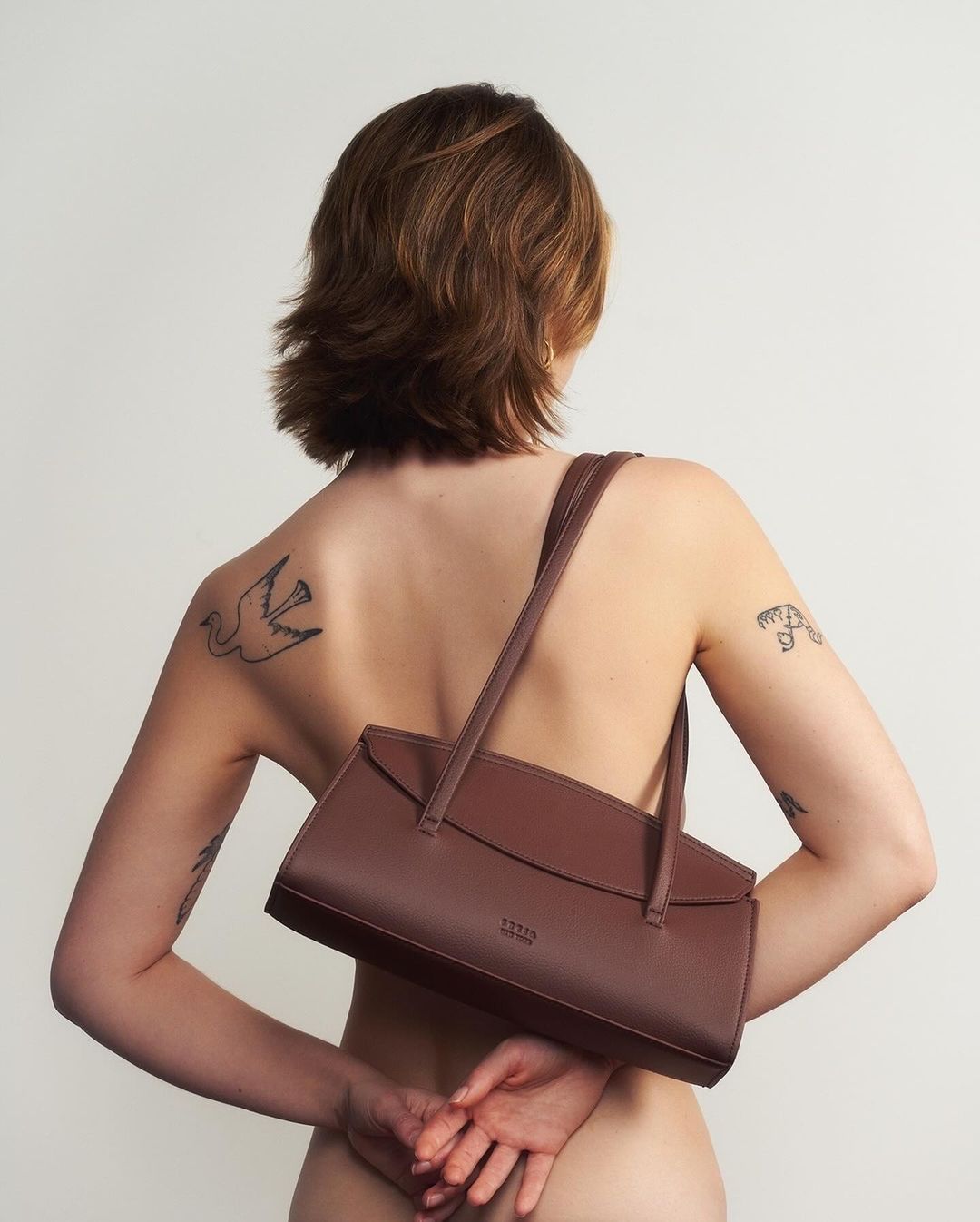 Woman poses from behind with brown purse over shoulder, tattoos on back and arms, in front of a light-colored wall.