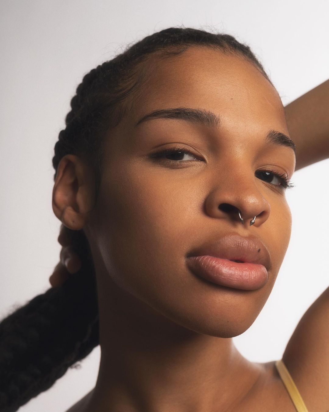 Close-up studio shot of a Black woman with braided hair and a septum piercing, posing against a white background with soft lighting.