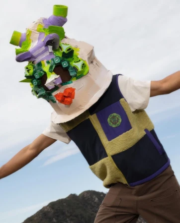 Person wearing a sculptural paper-mache mask and fleece vest against a sky background