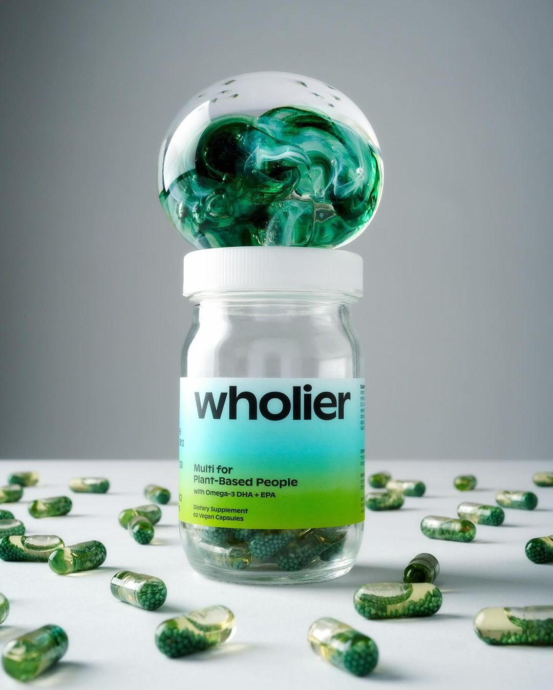 Close-up still life of a bottle of green-tinted multivitamin capsules, with loose capsules scattered around and a glass paperweight on top.