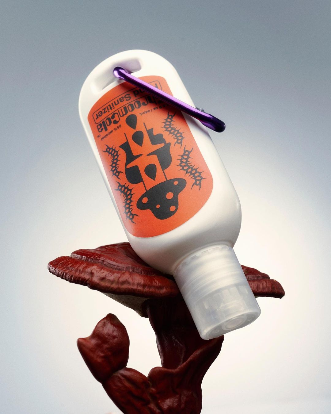 Hand sanitizer bottle with orange label and purple carabiner clip placed atop a reddish-brown ganoderma mushroom on a gradient gray background.