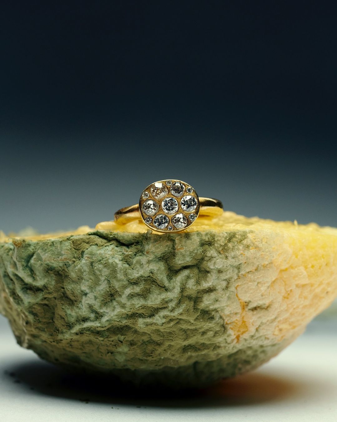 Close-up of a gold ring with a diamond cluster setting, displayed on a discolored, decaying citrus fruit against a blurred background.