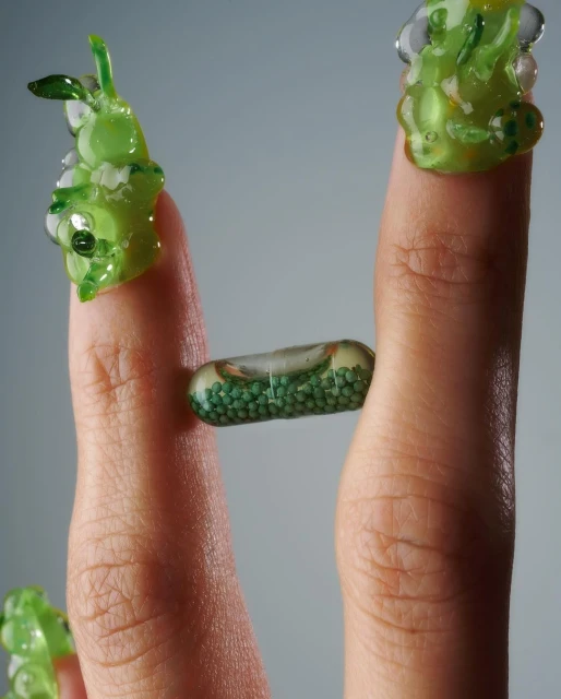 Woman's hand featuring creative nail art with transparent capsules filled with green beads and sculpted cartoonish green glass creatures.