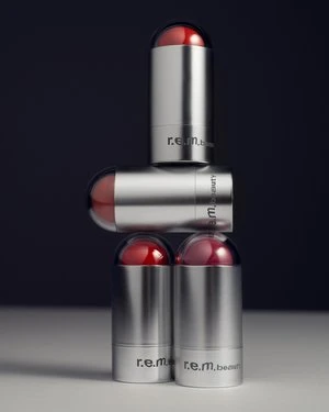 Stack of r.e.m. beauty lipsticks in silver tubes with red caps against a dark backdrop. Beauty product photography, close-up.