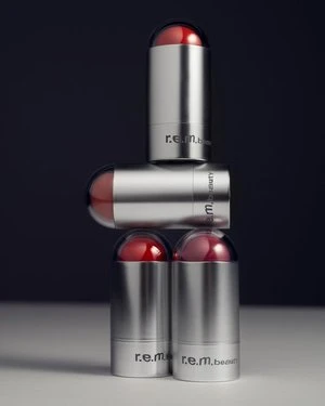 Product shot of a stack of three R.E.M. Beauty makeup products in silver tubes with red caps against a dark background.