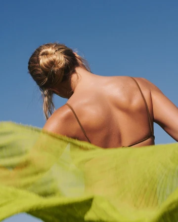 Woman seen from behind with blonde hair in a bun against a clear blue sky, partially obscured by a bright green flowing fabric