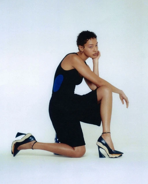 A slender woman with a short, curly haircut poses kneeling indoors wearing a black tank dress and stylish, heeled, two-tone dress shoes.