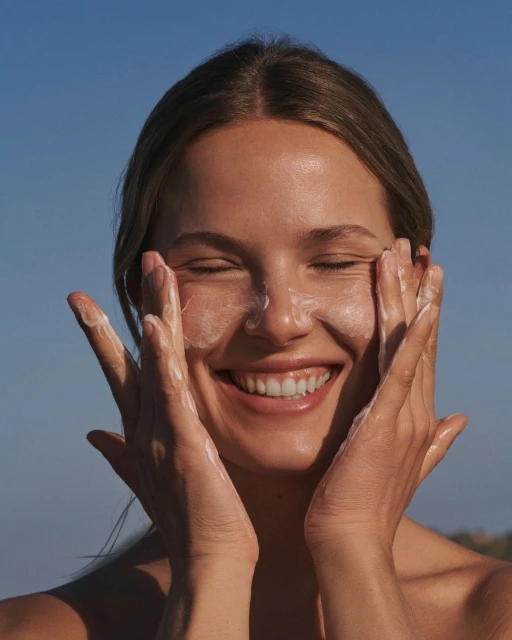 Happy young woman smiling and applying sunscreen to her face in an outdoor setting with a clear blue sky, skincare and sun protection.
