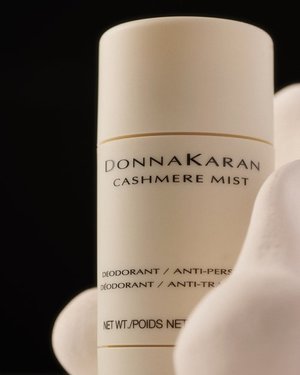 Donna Karan Cashmere Mist deodorant in a white container against a dark backdrop surrounded by soft, rounded shapes in a beauty product shot.