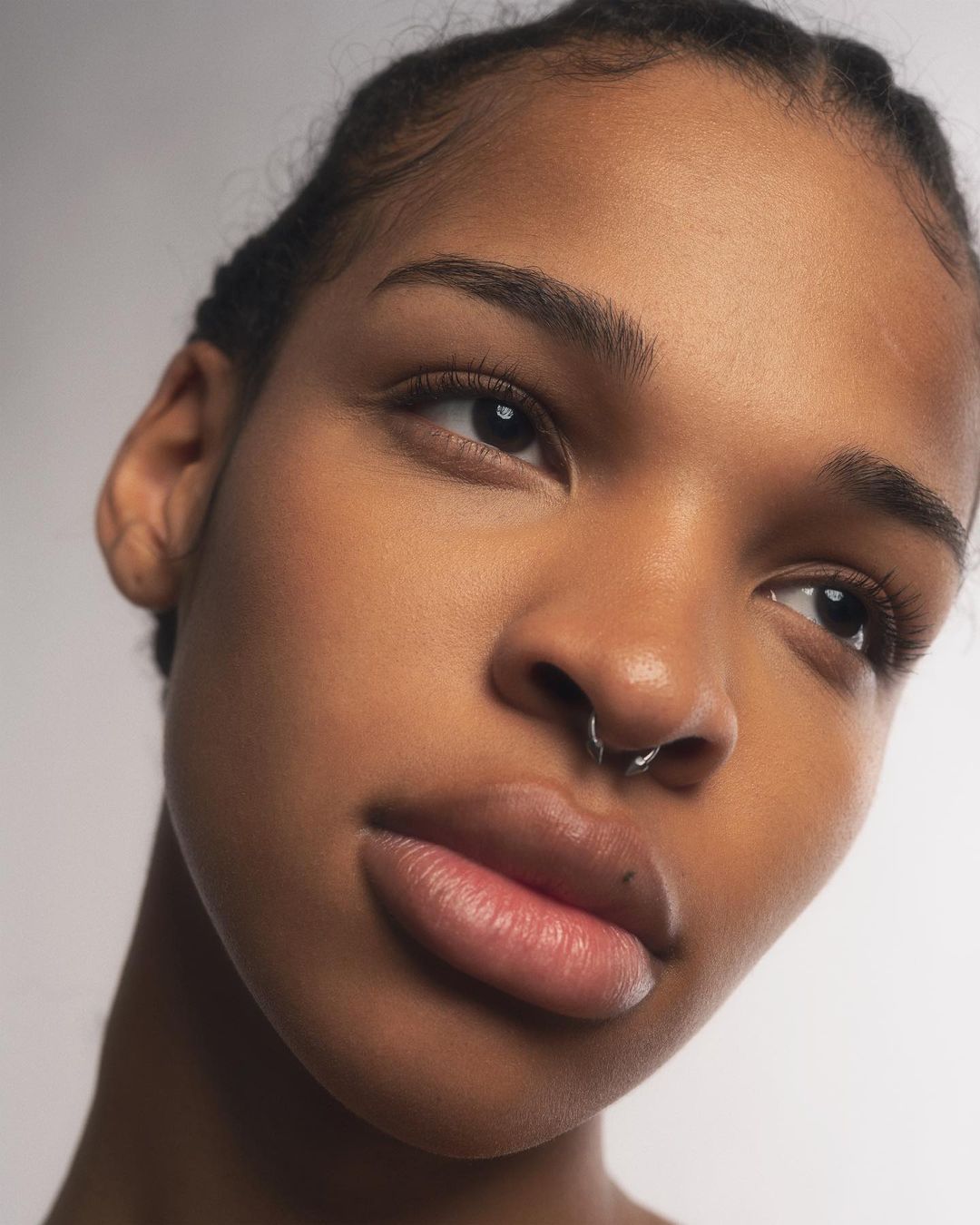 Close-up portrait of an adult woman with brown skin and dark hair, angled to the left, in a bright indoor setting against a neutral background.