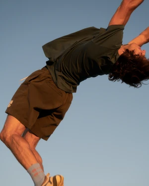Athletic man performs a backflip against a clear blue sky. He wears a dark green shirt and brown shorts in an outdoor fitness setting.