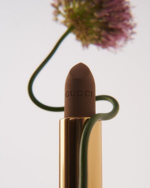 A close-up product shot features a brown Gucci lipstick centered with a purple allium flower and green stem against a white backdrop.
