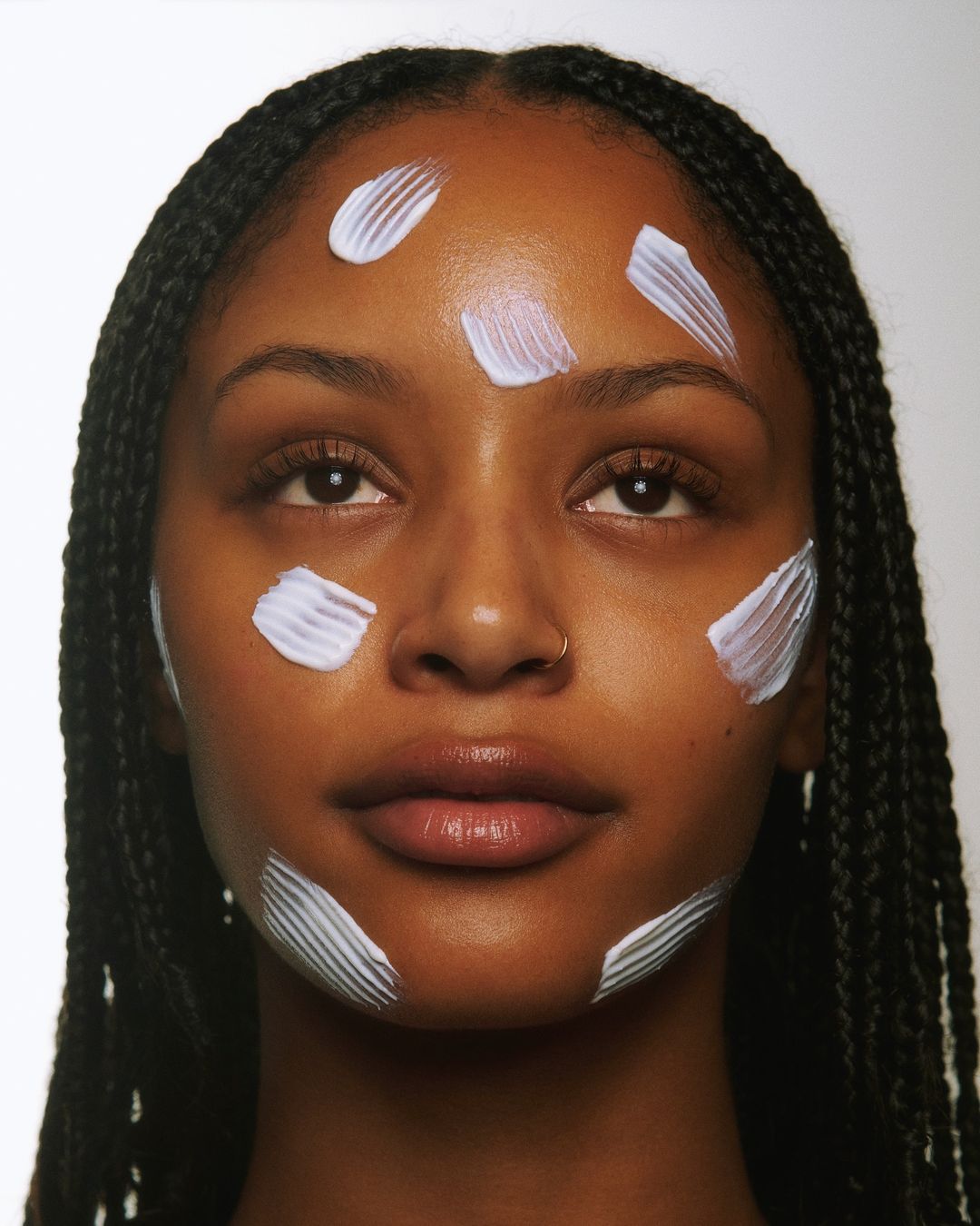 Close-up portrait of a woman with dark skin and braids, featuring streaks of white cream on her forehead, cheeks, and chin against white backdrop.