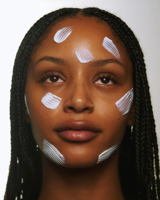 Close-up portrait of a woman with dark skin and braids, featuring streaks of white cream on her forehead, cheeks, and chin against white backdrop.