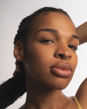 Close-up portrait of an African American woman with braided hair and a septum piercing posing in front of a neutral studio backdrop.