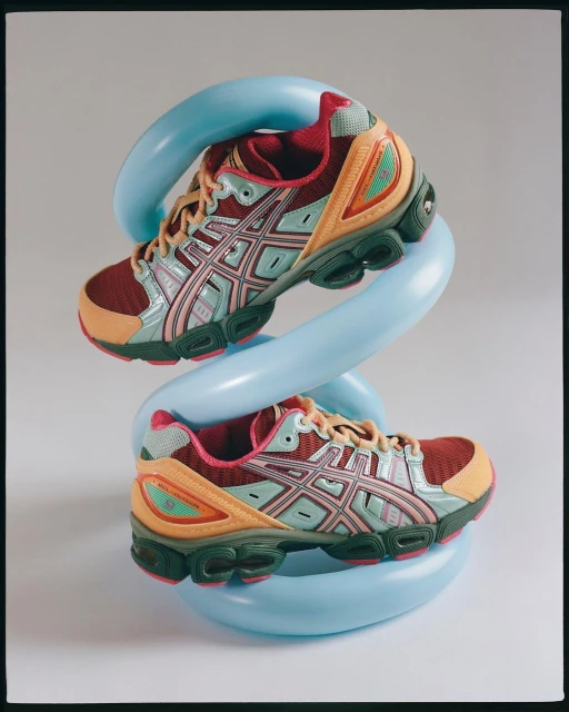 Two running shoes, one burgundy and one orange, intertwined with a light blue, twisty balloon on a white background in a studio.