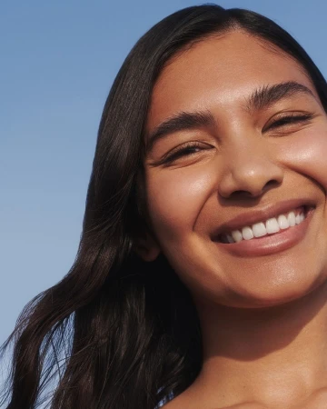 Close-up portrait of a smiling woman with dark hair against a clear blue sky background