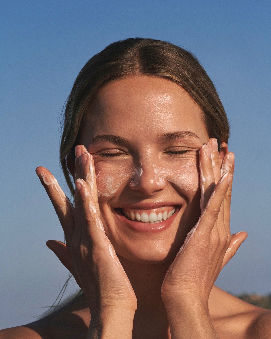 Close-up of a smiling young woman with eyes closed applying a creamy facial cleanser or moisturizer, outdoors against a blue sky.