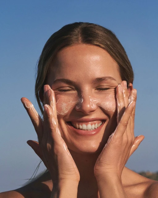 Close-up of a smiling young woman with eyes closed applying a creamy facial cleanser or moisturizer, outdoors against a blue sky.