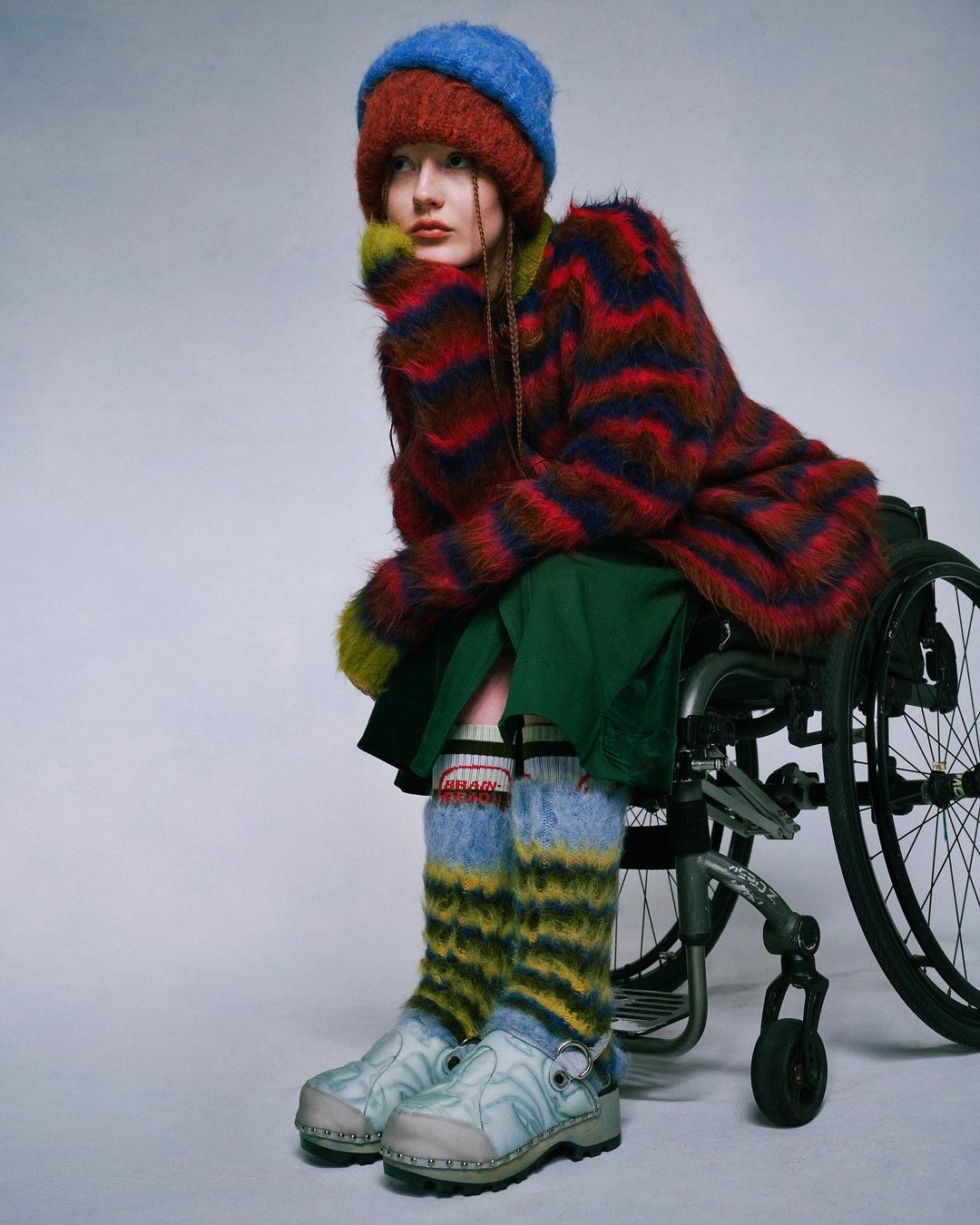 A young adult female model wearing colorful knitted hat, coat, and leg warmers poses in a wheelchair against a neutral background.