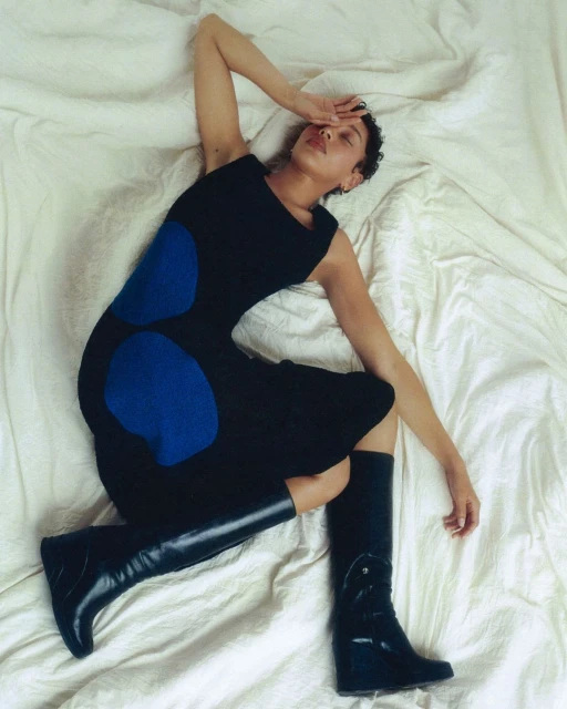Woman in blue and black dress and tall black boots poses on a crumpled white sheet. Arm covers her eyes.