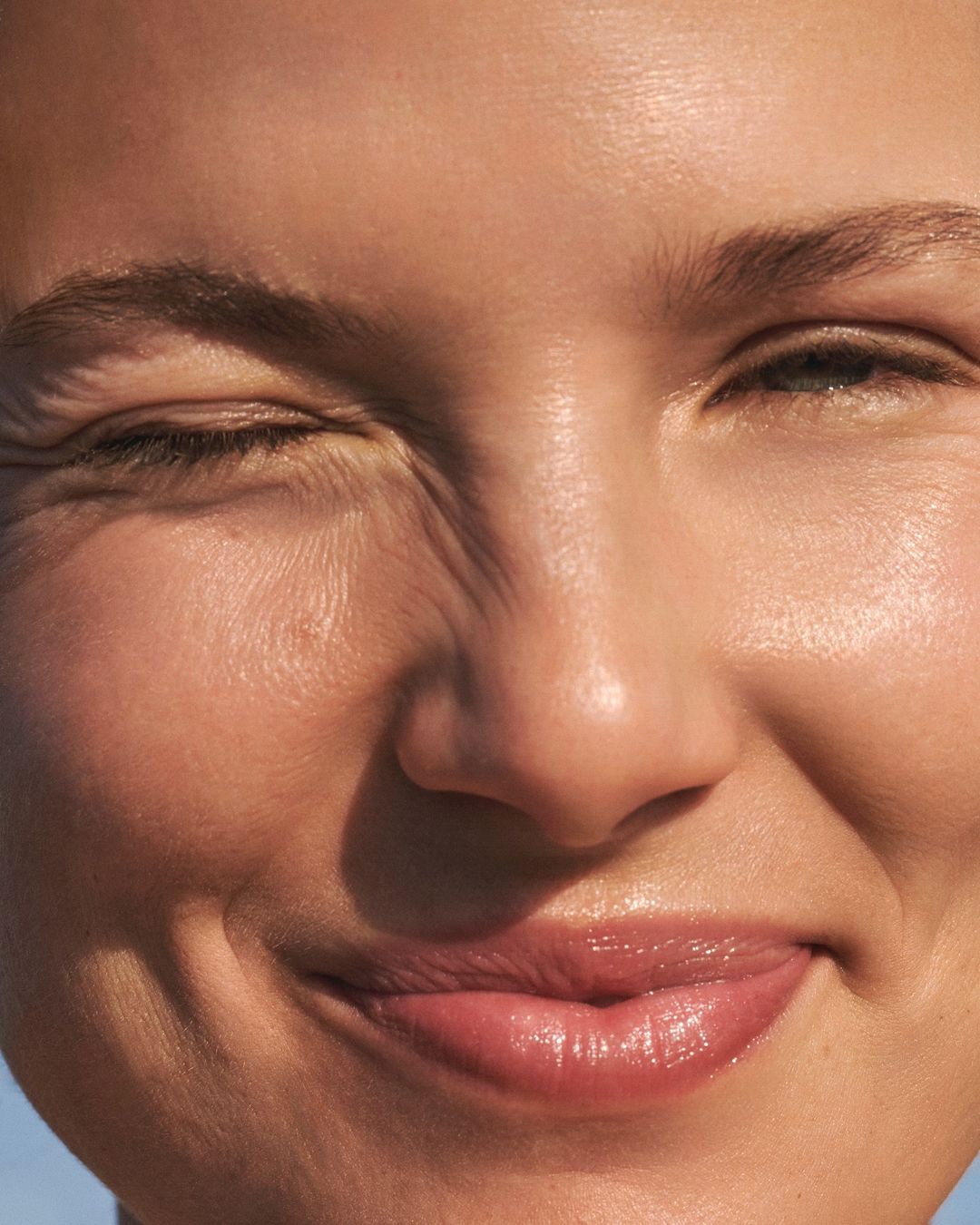 Close-up of a woman's face with sunlit, glowing skin, a slight smile, and one eye squinting, creating a sense of natural beauty and warmth.