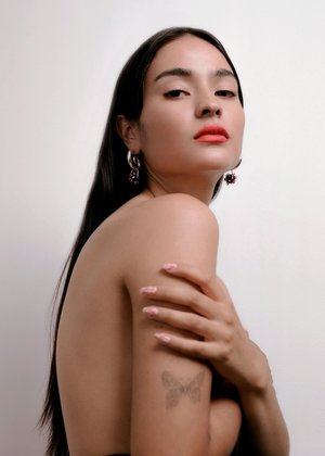 Portrait of a woman with long dark hair, wearing silver earrings, and coral lipstick against a plain white background in a close-up studio shot.