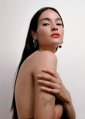 Portrait of a woman with long dark hair, wearing silver earrings, and coral lipstick against a plain white background in a close-up studio shot.