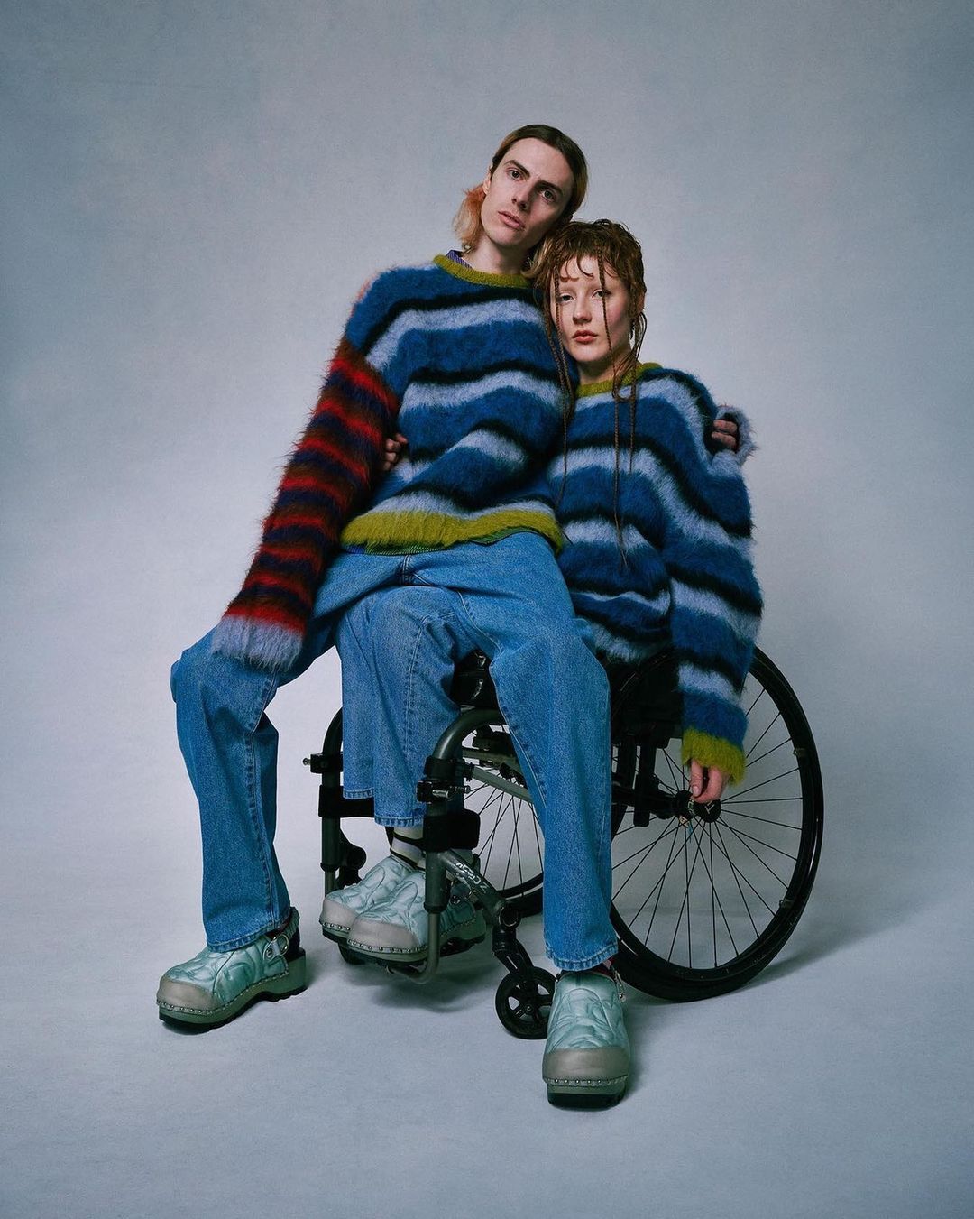 A stylish portrait showcases two people in striped sweaters. One sits in a wheelchair, embraced by the other, against a neutral backdrop.