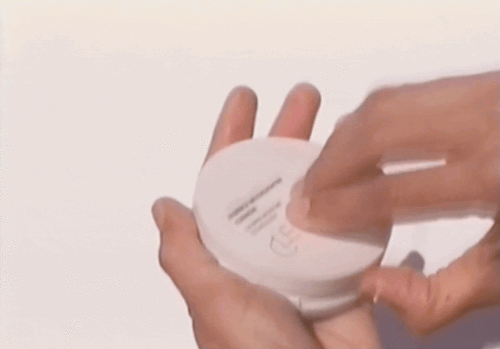 Close-up of hands opening a small round white cosmetic compact container against a plain background.