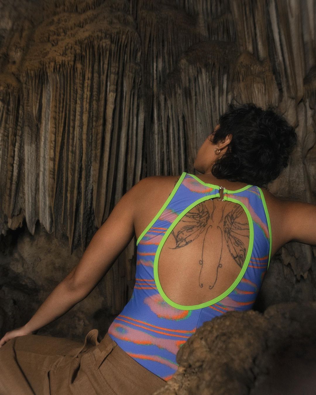Woman with a back tattoo wearing a blue and neon green swimsuit poses in a cave with stalactites in the background.