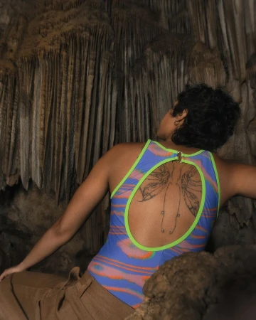 Woman with a back tattoo wearing a blue and neon green swimsuit poses in a cave with stalactites in the background.