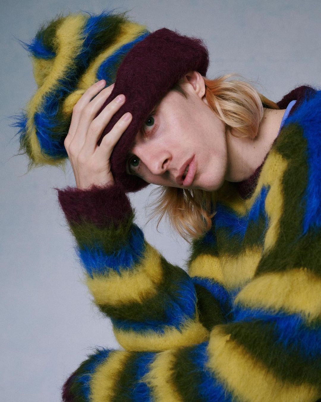 Portrait of a blonde male wearing a striped fur hat and sweater. The sweater features green, yellow, and blue horizontal stripes.