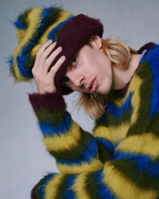 Portrait of a blonde male wearing a striped fur hat and sweater. The sweater features green, yellow, and blue horizontal stripes.