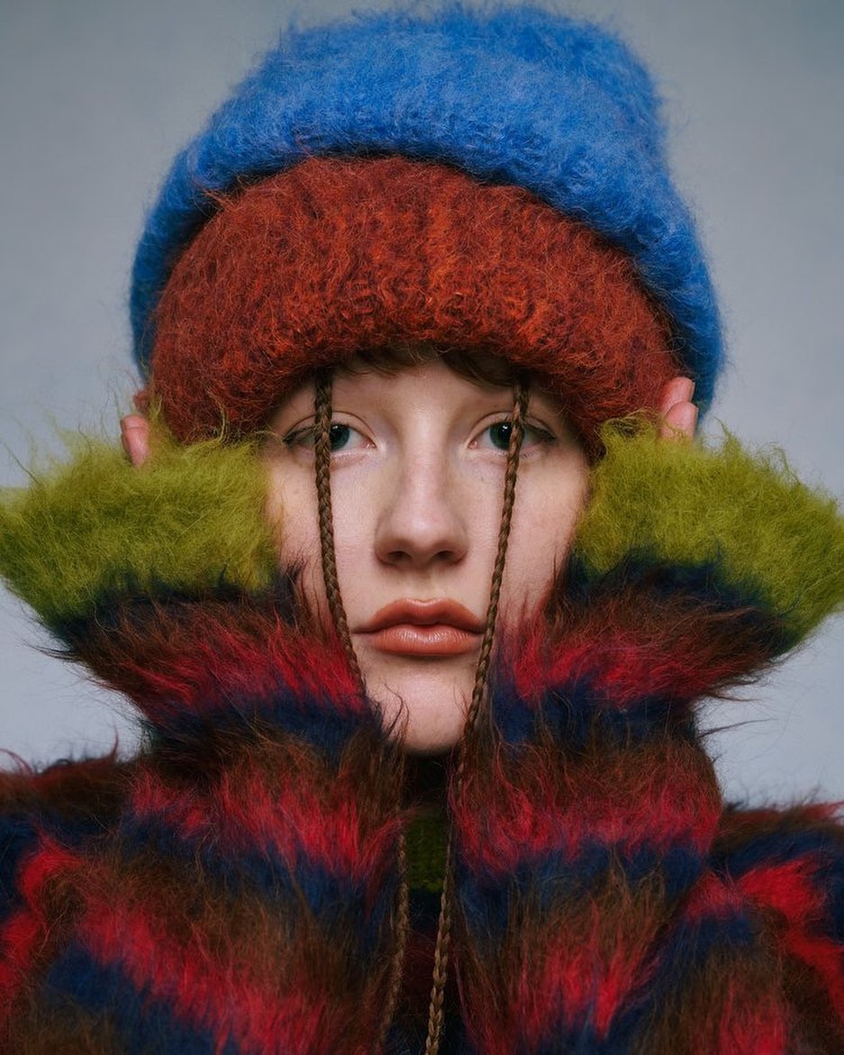 Close-up of a young woman wearing a triple-layered knit hat in blue, orange, and green, and a striped mohair jacket with braids.