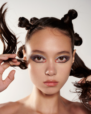 Close-up of a young adult female with styled dark brown hair and striking eyeshadow, posing against a plain white background.