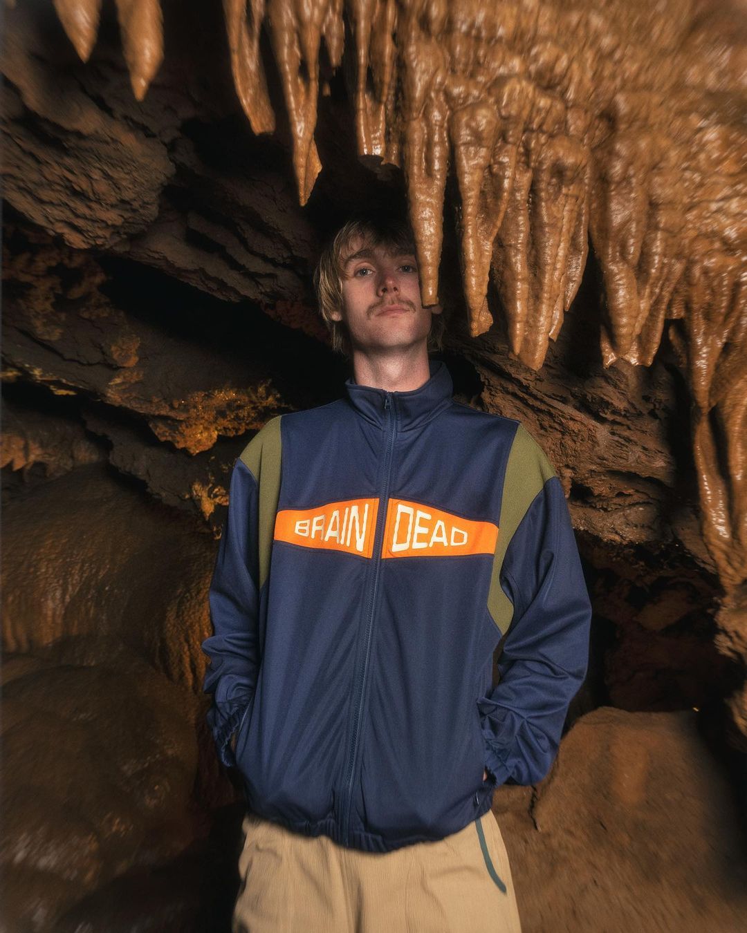Adult male posing in a cave wearing a Brain Dead jacket, tan pants, and with a visible mustache stands looking at the camera.