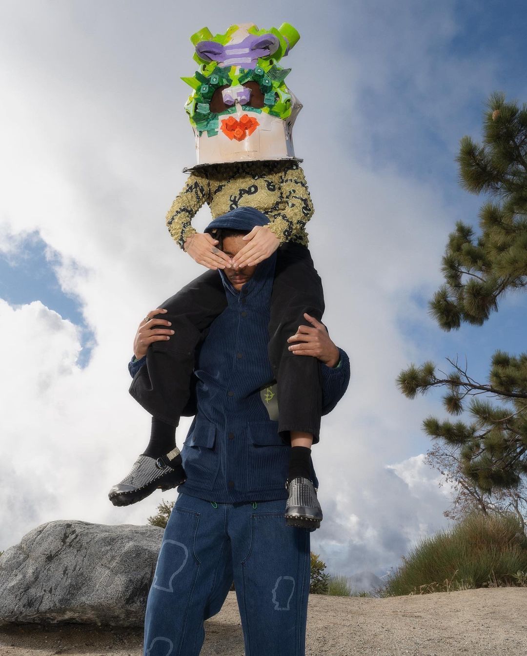 Two people outside, one wearing a decorated box on their head and sitting on the shoulders of another wearing blue jacket and pants.