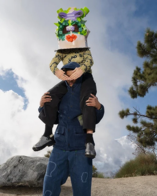 Two people outside, one wearing a decorated box on their head and sitting on the shoulders of another wearing blue jacket and pants.