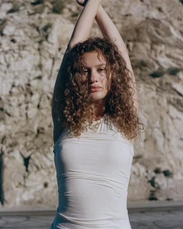 Woman with curly hair in a white top posing with arms raised against a rocky background photographed by Laviena Echev in Los Angeles.
