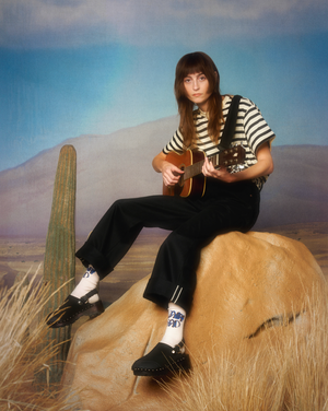 Woman in striped t-shirt and black trousers holding a guitar, seated on a painted desert backdrop with a cactus prop.