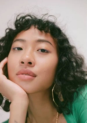 Close-up portrait of a young adult woman with curly dark hair and gold hoop earrings, resting her face on her hand in a relaxed pose.