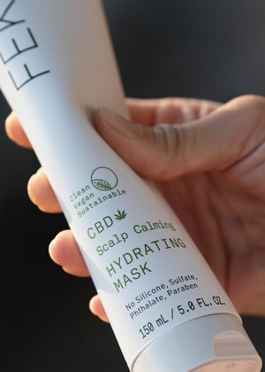 Close-up of a hand holding a white tube of CBD hydrating hair mask. Product claims include vegan, clean, and sustainable.