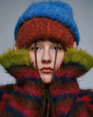 Portrait of a woman with fair skin, wearing colorful striped jacket and knit hats stacked on her head, against a neutral backdrop.