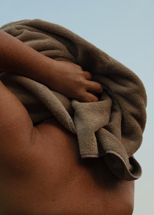 Close-up of a person drying their skin with a beige towel, water droplets visible, against a light blue sky background.