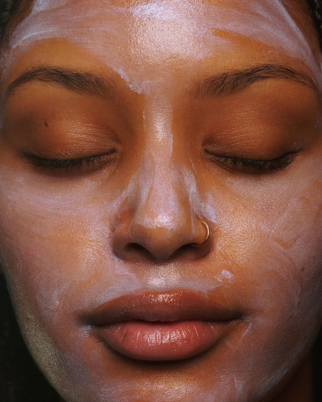 Close-up studio shot of a Black woman with her eyes closed, wearing a facial mask, and a nose ring, showcasing her skincare routine.